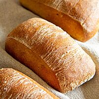 Drei ausgebackene Weizenmischbrötchen liegen mit glatter, leicht bemehlter Kruste in einem Leinentuch.
