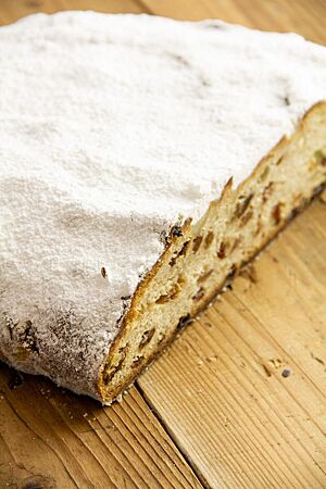 Ein flacher, leicht ovaler Stollen mit einer dicken Schicht Puderzucker liegt auf einem hellbraunen Holztisch.