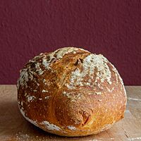Ein kräftig ausgebackenes Weizenbrot mit leicht bemehlter Kruste liegt auf einem Holztisch.