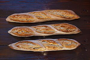 Drei an den Enden spitz zulaufende Altbrotstangen mit leicht bemehlter Kruste liegen auf einem Holztisch.
