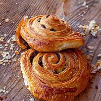 Zwei goldbraun ausgebackene Rosinenschnecken liegen auf einem Holztisch.