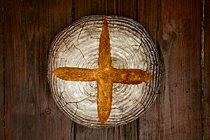 Auf einem Holztisch liegt kreuzförmig eingeschnitten ein kräftig ausgebackenes Landbrot.