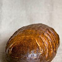 Das dunkel ausgebackene Dreierbrot mit glänzender Kruste liegt auf einem Leinentuch.