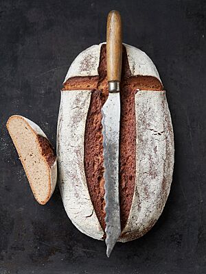 Auf dem länglich geformten, kräftig ausgebackenen Rolandbrot mit bemehlter und kreuzförmig eingeschnittener Kruste liegt ein langes, antikes Wellenschliffmesser.