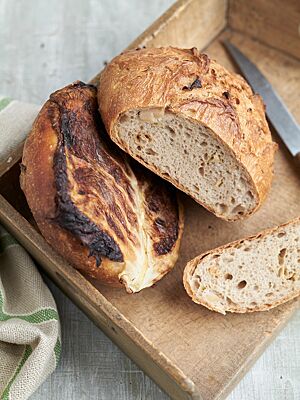 Ein Kohlbrot mit stark gebräuntem Weißkohlblatt und ein Kohlbrot mit rustikal aufgerissener Kruste liegen auf einem Holzbrett. In der lockeren Krume des Dithmarscher Kohlbrotes sind Weißkohlstücke zu erkennen.