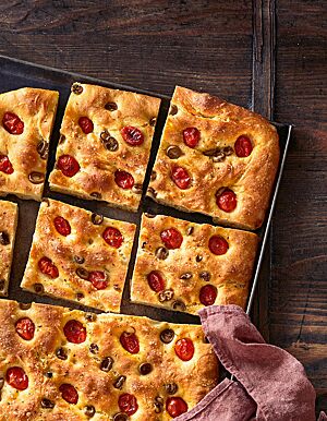 Die saftige Focaccia mit Tomaten, Oliven und Salz liegt geschnitten auf einem Backblech.