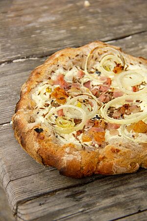 Der mit Schmand, Zwiebeln und Speck belegte Hitzplootz liegt fertig ausgebacken auf einem rustikalen Holztisch.
