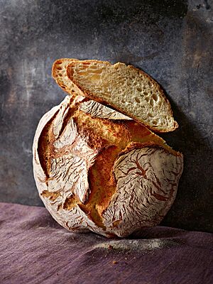 Der goldbraun ausgebackene, runde Laib des Weizenbrotes hat eine rustikal aufgerissene Kruste und eine mittelporige, hellgelbe Krume.