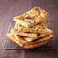 Mehrere Stücke Flammkuchen liegen gestapelt auf einem Gitter, sodass der verhältnismäßig dicke, fluffige Boden gut sichtbar ist.