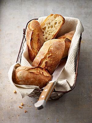 Längliche, rustikal aufgerissene Baguettebrötchen mit grobporiger Krume liegen in einem Brötchenkorb.