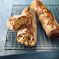 Zwei goldbraun ausgebackene und der Länge nach mittig aufgerissene Baguettes mit grobporiger, heller Krume liegen auf einem Rost.