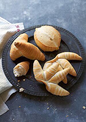 Zwei goldgelb ausgebackene Fingerbrötchen mit gewickelter Struktur und glänzender, glatter Oberfläche (rechts) liegen mit einem Mailänder Brötchen (Mitte) und zwei Cornetti (links) auf einer dunklen Platte.