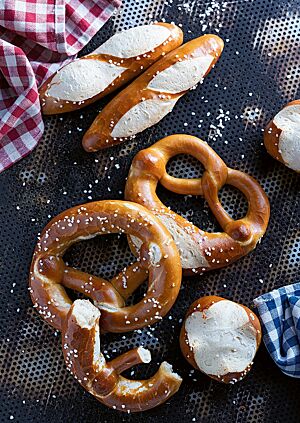 Auf einem dunklen Backblech mit Lochung liegen Laugenbrezeln (Mitte), Laugenstangen (oben) und Laugenbrötchen (unten). Ihre glänzende, feine Kruste ist dunkelbraun ausgebacken und mit grobem Salz bestreut.
