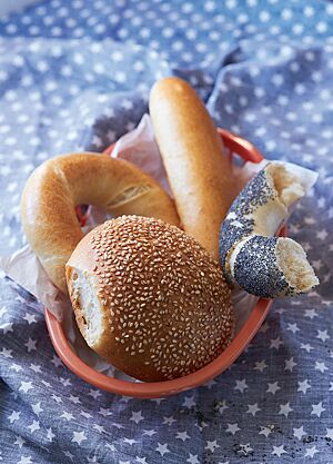 Auf einer blauen Decke mit weißen Sternen steht ein Brotkorb, der mit einem Sandwich-Brötchen (oben), eineinhalb Bagels (links und rechts) und einem Hamburger Bun (unten) gefüllt ist. Das Sandwich-Brötchen ist lang, schmal und hat eine stark glänzende, goldgelb ausgebackene, feine Kruste.