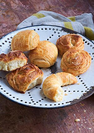 Auf einer weißen Emailleplatte liegen Splitterhörnchen (unten rechts), Dänische Brötchen (oben links) und Splitterbrötchen in der Ost- und Westvariante (unten links und oben rechts). Die leicht eckigen Splitterbrötchen (Ost) haben eine wild-blättrige, goldbraun ausgebackene Kruste, die an eine aufblühende Knospe erinnert.