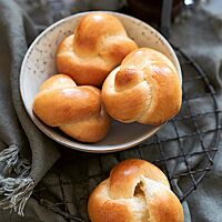 Vor und in einer hellen Schale liegen drei Zopfknoten (links und unten) und ein Zopfknopf (rechts). Die Kruste der Zöpfe ist goldgelb ausgebacken und glänzt stark.