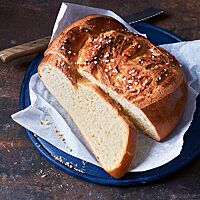 Das angeschnittene Campingbrot liegt auf Pergamentpapier auf einer blauen Platte. Die helle Krume ist wattig-weich, die rustikale Kruste goldbraun ausgebacken. Im Ausbund des Brotes kommt der eingearbeitete Hagelzucker zum Vorschein.