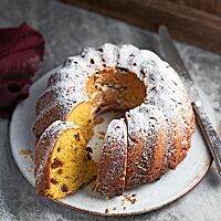 Der mit Puderzucker bestäubte Gugelhupf liegt auf einem weißen Teller. Der Anschnitt lässt die gelbe, mit Sultaninen durchzogene Krume erkennen.