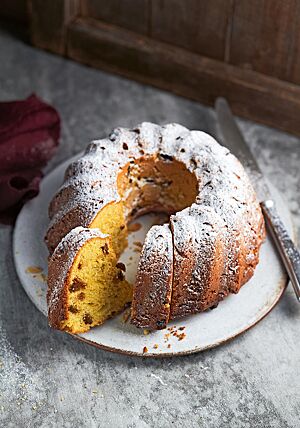 Der mit Puderzucker bestäubte Gugelhupf liegt auf einem weißen Teller. Der Anschnitt lässt die gelbe, mit Sultaninen durchzogene Krume erkennen.
