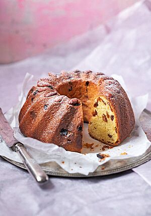 Der angeschnittene französische Gugelhupf liegt auf einem Pergamentpapier auf einem Silberteller. Die mit Rosinen durchzogene Krume ist feinporig und faserig. Auf jeder Wölbung des Gugelhupfes liegt oben eine eingebackene Mandel.