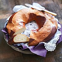 Eine ringförmige Pogne de Romans mit stark glänzender Kruste liegt angeschnitten auf einem weißen Spitzendeckchen. Die wattig-weiche Krume hat eine kleine, regelmäßige Porung.