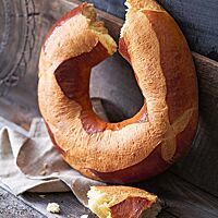 Eine ringförmige Pogne de Romans lehnt senkrecht an einer Holzwand. Im oberen Teil des Ringes ist ein Stück herausgebrochen, das vor dem Brot auf dem Holzuntergrund liegt. Die wattig-weiche, helle Krume wird von einer dünnen, goldbraunen Kruste umhüllt.