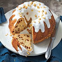 Ein angeschnittener polnischer Babka mit einer dicken Schicht Zuckerguss und gehackten Walnüssen auf der Oberfläche liegt auf einer weißen Platte. Im Anschnitt sind viele Sultaninen in der elastischen, saftigen Krume erkennbar.