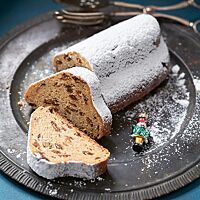 Der mit Puderzucker bestreute Quarkstollen liegt auf einer dunklen, runden Platte. Zwei abgeschnittene Scheiben liegen vor dem Stollen. Sie zeigen die saftige, feinporige Krume.
