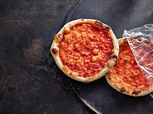 Zwei nur mit Tomatensauce bestrichene Pizzen weisen am Rand der hellen Kruste einige dunkle Stellen auf.