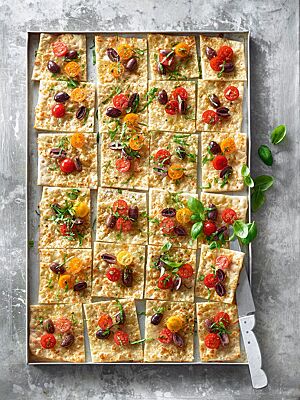 Auf einem Blech liegt die mit Mozzarella, Kirschtomaten, Oliven und Basilikum belegte rechteckige Pizza Romana.