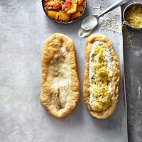 Zwei goldgelb frittierte, längliche Lángos liegen mit einer Schüssel Letscho auf einem hellen Blech: ein Lángos bestrichen mit Knoblauchwasser (links) und eines belegt mit Sauerrahm und geriebenem Käse (rechts).