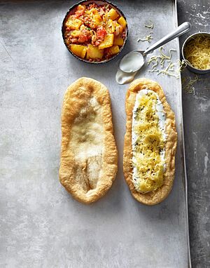 Zwei goldgelb frittierte, längliche Lángos liegen mit einer Schüssel Letscho auf einem hellen Blech: ein Lángos bestrichen mit Knoblauchwasser (links) und eines belegt mit Sauerrahm und geriebenem Käse (rechts).