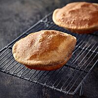 Zwei runde, aufgeblasene Puri liegen auf einem eckigen Gitterrost. Sie wurden goldbraun und knusprig frittiert.