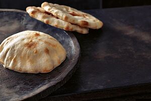 Zwei flache Pitas liegen angelehnt an den Rand eines Teller, auf dem sich eine aufgeblähte Pita befindet. Die kleinen Fladenbrote haben eine helle Krume mit einigen braunen Stellen.