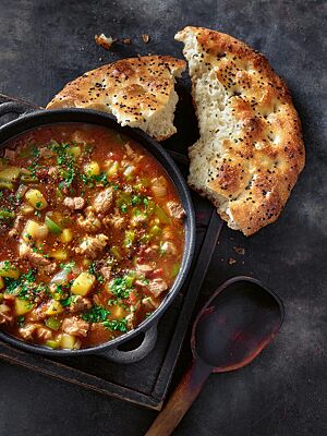 Neben einem Topf mit orientalischer Gulaschsuppe liegt eine große angebrochene Pita mit fluffiger, offenporiger Krume und goldgelb bis goldbraun ausgebackener Kruste.