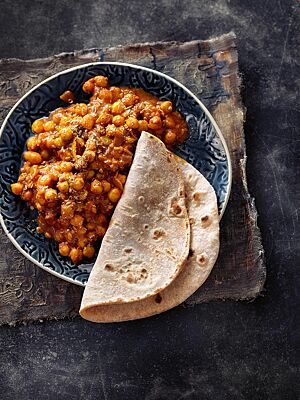 Ein Chapati liegt auf dem Rand einer Schale mit Channah Masala. Das dünne, runde Fladenbrot ist hell ausgebacken mit einigen dunklen Stellen.