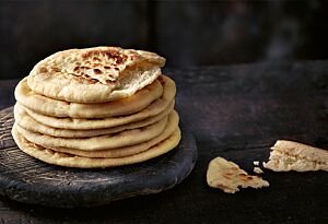 Ein Stapel Pane ’e Poddine liegt auf einem runden Holzbrett. Die sardischen Fladenbrote sind hell ausgebacken mit einigen dunklen Stellen und innen hohl, da sie sich beim Backen aufgebläht haben.