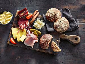 Kräftig ausgebackene Paarlen liegen zusammen mit Schinken, Käse, Speck, Würstchen, Tomaten, Gewürzgurken und einem Schälchen geriebenem Meerrettich auf einem großen Holzbrett. Die paarweise aneinandergebackenen Paarlen haben eine glatte, bemehlte Kruste und eine weiche, lockere Krume.