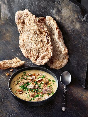 Zwei flache, durchgetrocknete Schüttelbrote lehnen aufrecht an einer Wand neben einer Schüssel Knoblauchrahmsuppe mit Schüttelbrotstückchen.