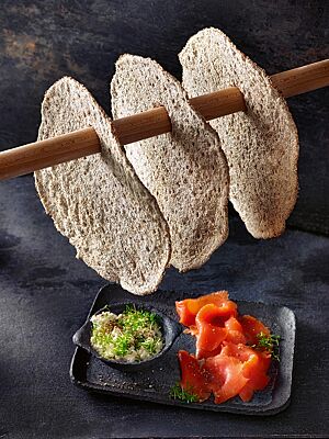 Auf einer kleinen Platte liegen Räucherlachs und mit Kresse bestreuter körniger Frischkäse. Darüber hängt eine Stange, an der drei dünne, runde Knäckebrote aufgehängt sind.