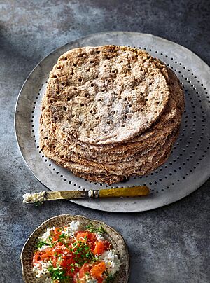 Auf einer runden Platte liegen neben einem Schälchen mit körnigem Frischkäse und Tomaten übereinandergestapelt mehrere runde Flatkaka mit gestippter, leicht bemehlter Oberfläche.