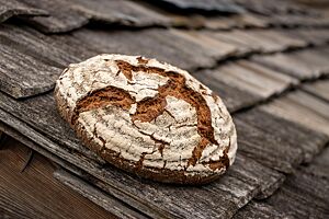 Der runde Laib des kräftig ausgebackenen Roggenbrotes hat eine bemehlte Kruste und liegt auf verwitterten Dachschindeln aus Holz.
