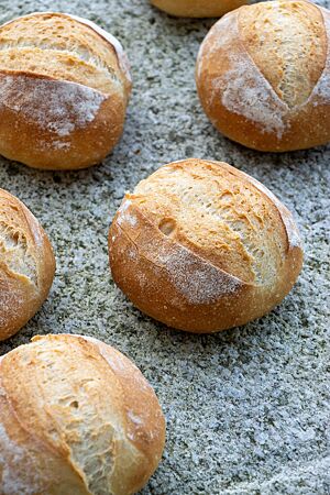 Knusprige, goldbraun ausgebackene Weizenbrötchen liegen auf einer Steinplatte.