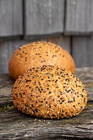Zwei goldgelb ausgebackene Roggenbuns mit Sesam auf der Oberfläche liegen auf einem rustikalen Holztisch.