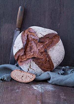 Das runde Weizenmischbrot hat eine bemehlte Kruste mit einem dunkel ausgebackenen, kreuzförmigen Einschnitt. Der Laib liegt auf einem Tuch, angelehnt an eine Holzwand. Die Krume des Brotes ist elastisch und locker.