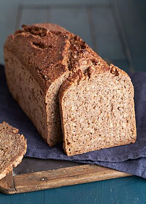 Der angeschnittene, in der Form gebackene Feldhamster liegt auf einem dunkelblauen Tuch. In der an den Laib gelehnten Brotscheibe sind die Roggenkörner in der Krume deutlich sichtbar. Die glänzende Kruste ist rustikal aufgerissen.