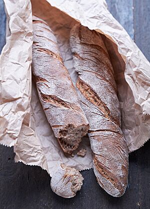 Die beiden Knorrigen mit bemehlter, rustikaler Kruste liegen in einer aufgerissenen, braunen Papiertüte. Das abgebrochene Stück Brot lässt die kleinporige, elastische Krume sichtbar werden