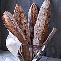 Drei Pain de Luxe-Stangenbrote und ein Brotmesser stecken aufrecht in einem mit einer weißen Serviette ausgelegten Metallkorb. Im Längsschnitt eines Brotes wird die unregelmäßige, wilde Porung der saftigen Krume sichtbar.