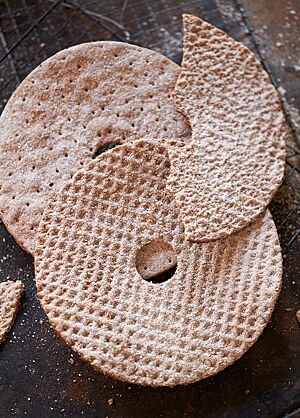Auf einem Gitterrost, der auf einem dunklen Untergrund steht, liegen mehrere runde Scheiben Knäcke mit einem Loch in der Mitte. Wie das Muster des Brotes erkennen lässt wurden die Scheiben vor dem Backen mit einer Knäckebrotrolle bzw. einer Stipprolle bearbeitet.