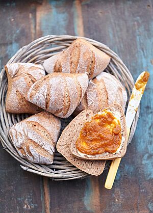 Die eckigen Weizenkissen mit bemehlter, mehrfach parallel eingeschnittener Kruste liegen in einem Holzkorb. Ein aufgeschnittenes Brötchen wurde mit einem orangenfarbenen Fruchtaufstrich bestrichen. Die Krume der Brötchen ist elastisch und kleinporig.
